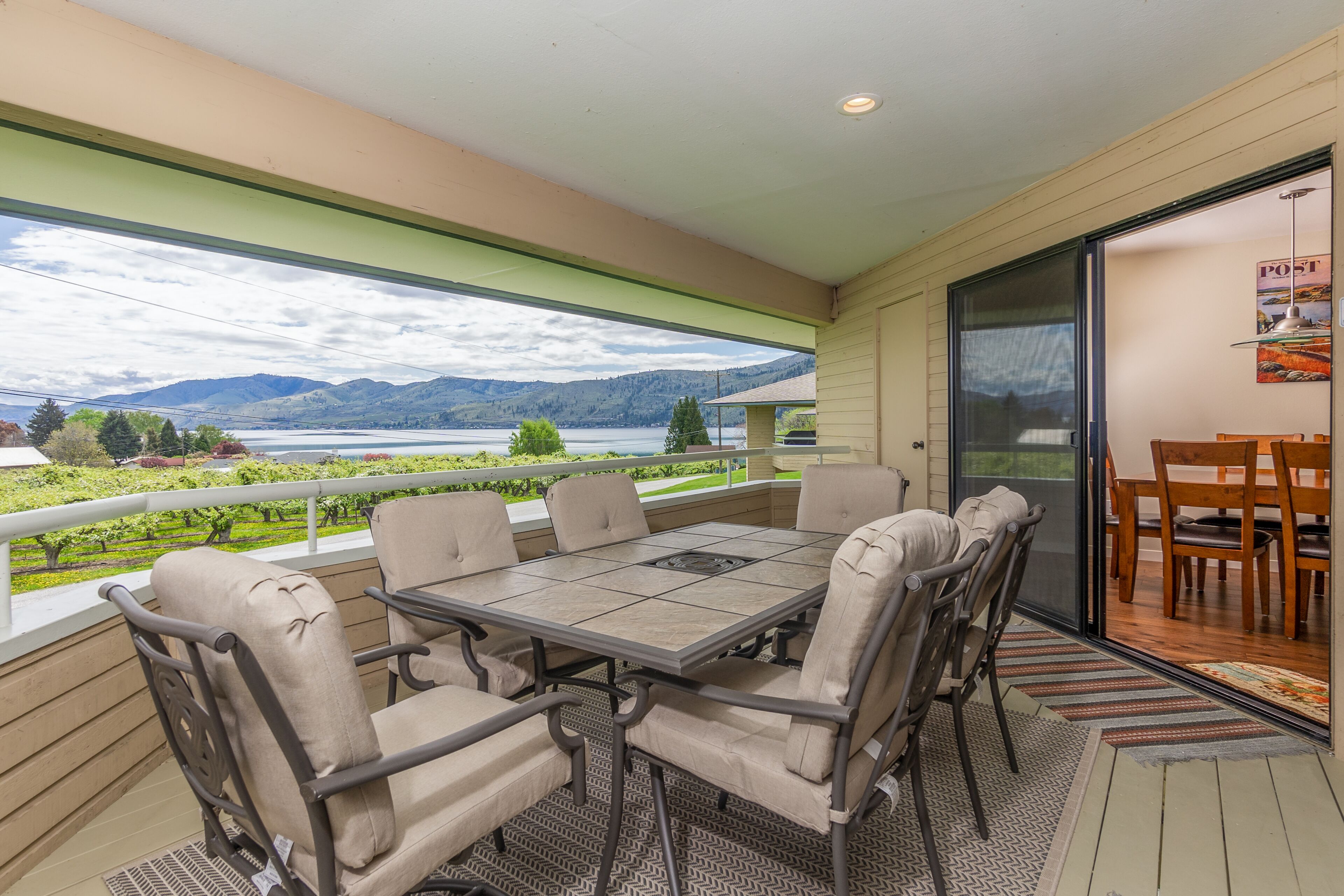 Un balcón con una mesa y sillas, vista a un lago y montañas, y una puerta que da a otra habitación.