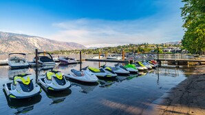 A marina with several jet skis docked at the pier, a dock, and a backdrop of mountains and a residential area.