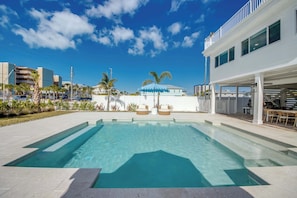 A swimming pool with a clear blue water, surrounded by a paved area, and a building with a balcony and outdoor seating.