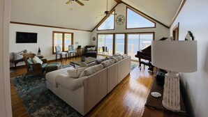 A spacious living room with a high ceiling, large windows, a piano, and a cozy seating area. - Your Private Coastal NC Waterfront Escape. Dock, deck, sun, peace, REPEAT! (Aurora)