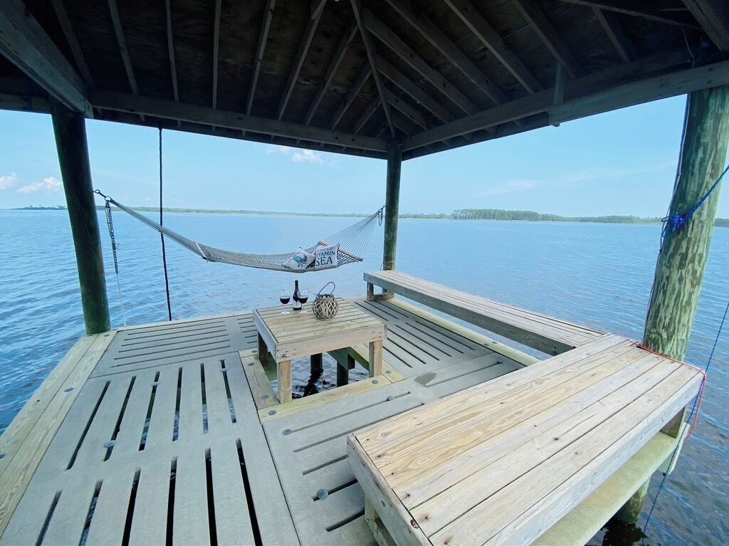 A wooden deck with a hammock, a table with a bottle, and a view of the water.