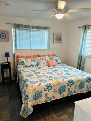 Un dormitorio con cama, ventilador de techo, mesita de noche, lámpara y ventana con cortinas.
