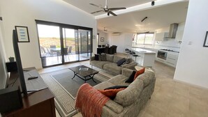 A spacious living area with a large sofa, a coffee table, and a TV. There is a sliding glass door leading to an outdoor patio. The kitchen is visible in the background with modern appliances. - 2 Corella Court - Private Jetty for a boat up to 12mt. (EXMOUTH)