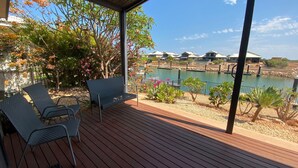 A covered outdoor area with a wooden deck, two lounge chairs, and a view of a swimming pool and houses in the background. - 2 Corella Court - Private Jetty for a boat up to 12mt. (EXMOUTH)