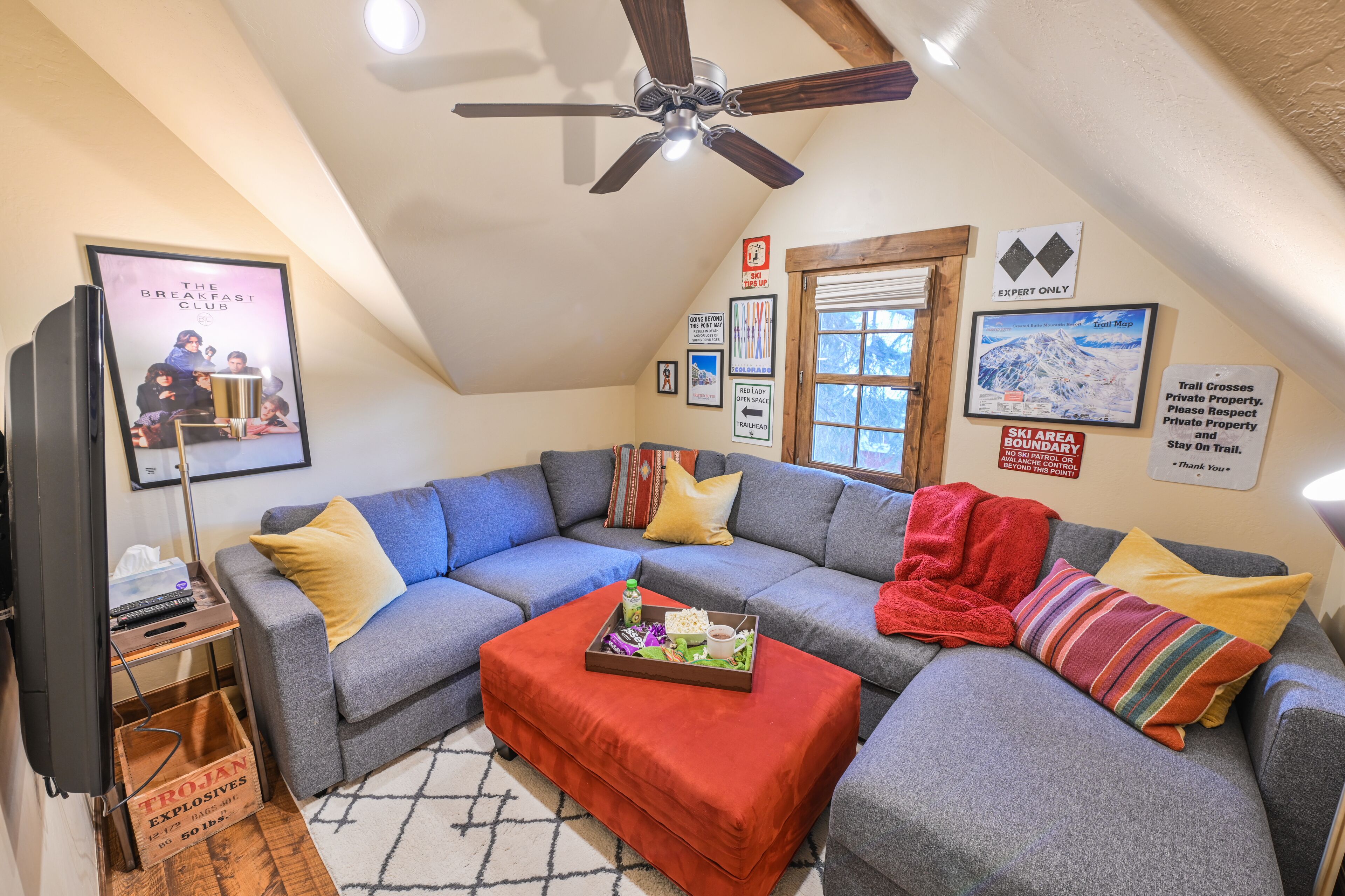 Un salon chaleureux avec un canapé d’angle, un pouf rouge et un ventilateur de plafond.
