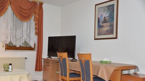 Huoneessa on puinen työpöytä, kaksi tuolia, televisio, kehystetty taulu ja ikkuna, jossa on verhot.