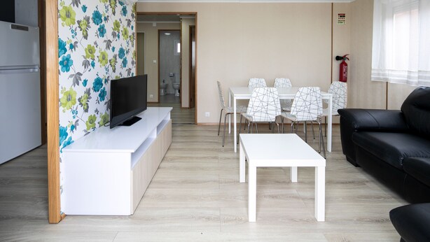 Una sala moderna con una televisión de pantalla plana, una mesa de comedor con sillas, un sofá de cuero negro y una cocina con refrigerador.
