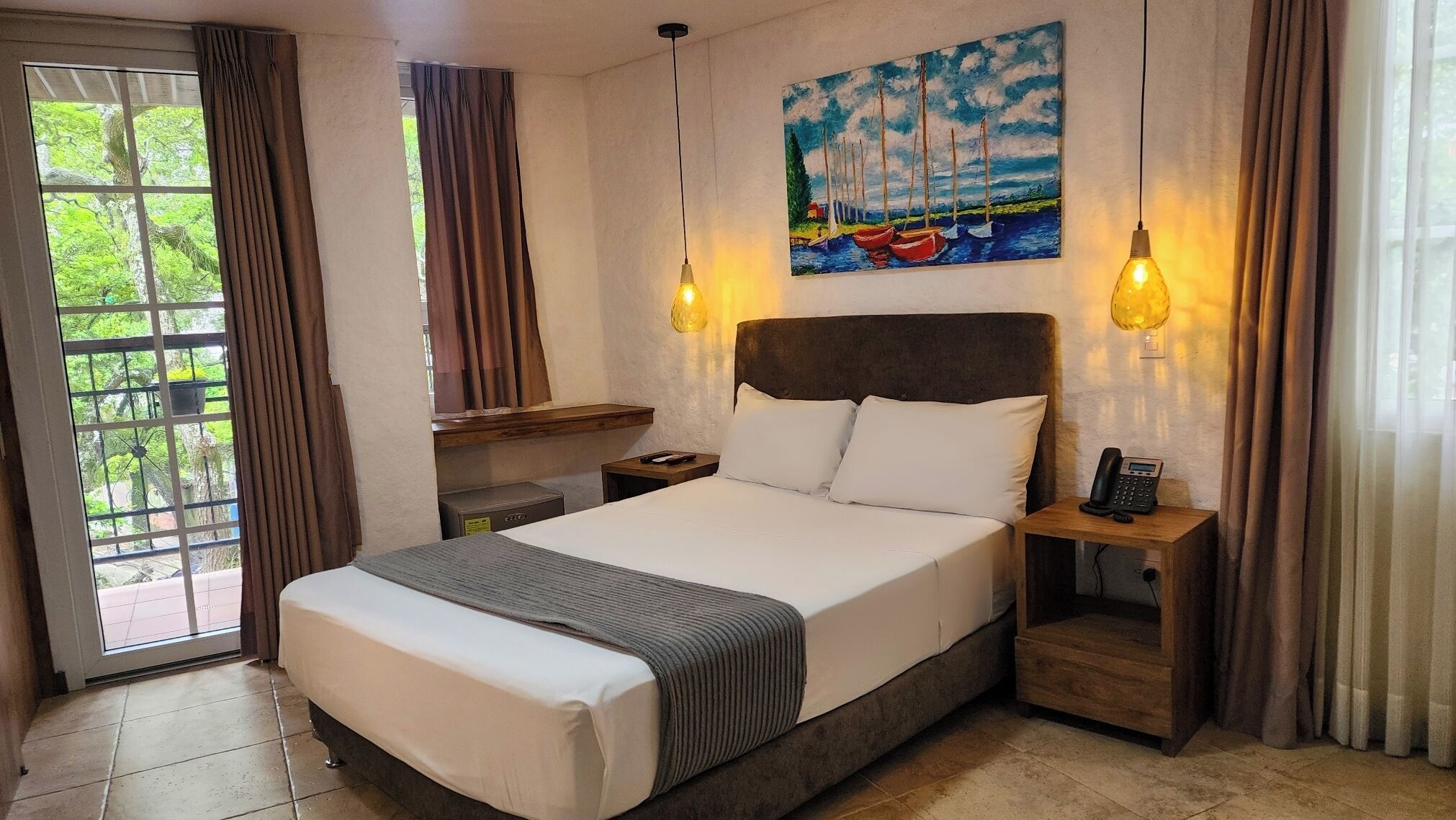 Ein Hotelzimmer mit einem großen Bett, zwei Nachttischlampen, einem Bild von Booten und Blick durch ein Fenster.