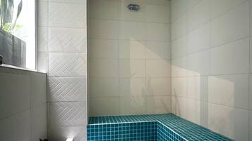Un baño con paredes blancas, piso de baldosas, un lavamanos en esquina y un banco azulado de azulejos.