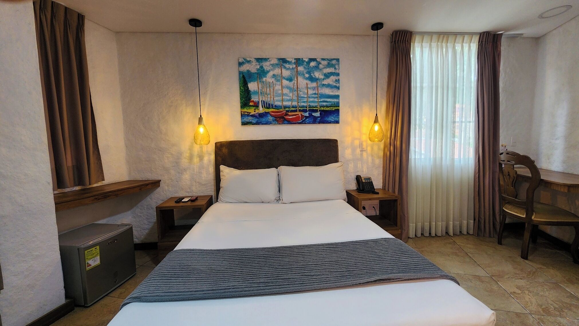 Habitación de hotel con cama, dos mesitas de noche, escritorio, silla, un cuadro y cortinas.