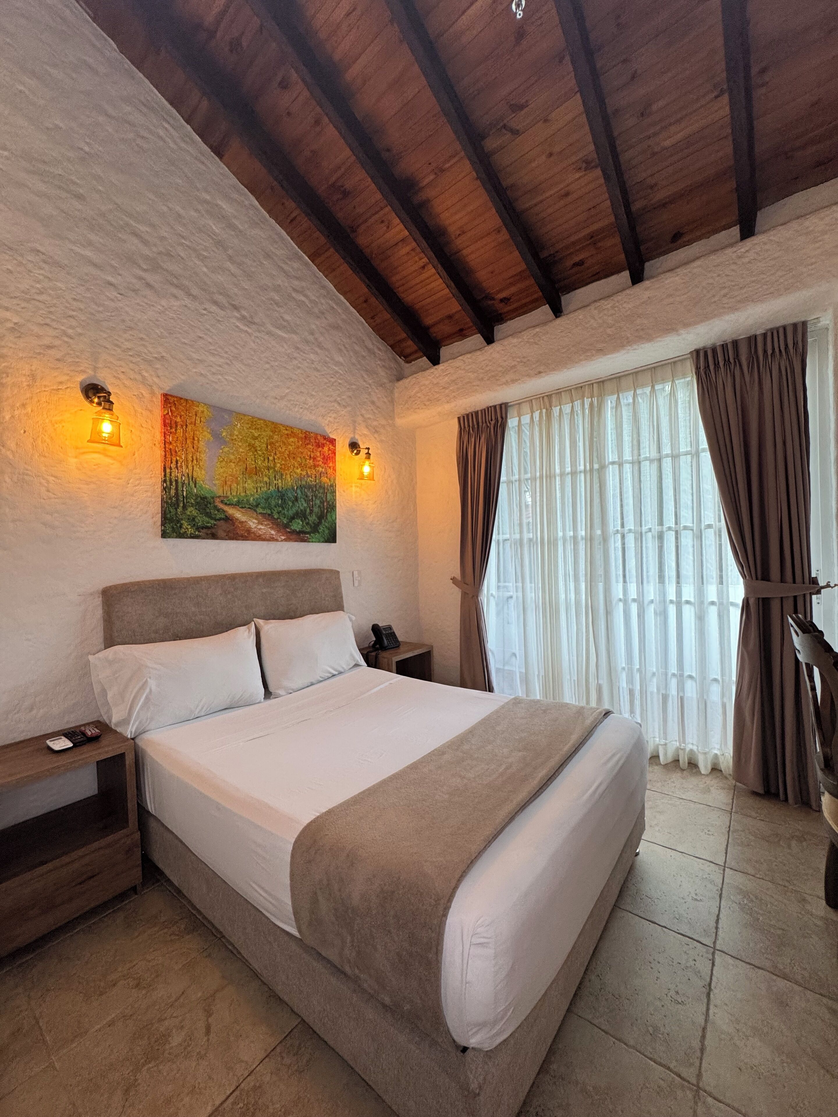 Quarto com cama grande, vigas de madeira no teto, um quadro na parede e uma janela com cortinas.