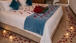 Un dormitorio con cama, almohadas, una manta azul, un ramo de rosas rojas y pétalos de rosa esparcidos.
