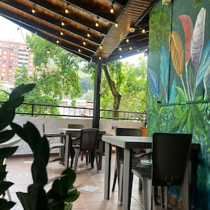 An outdoor cafe area with tables and chairs, a wooden ceiling with hanging lights, and a mural of flowers on the wall. - Hotel Casa del Hidalgo Centenario - Business & Leisure (Cali)