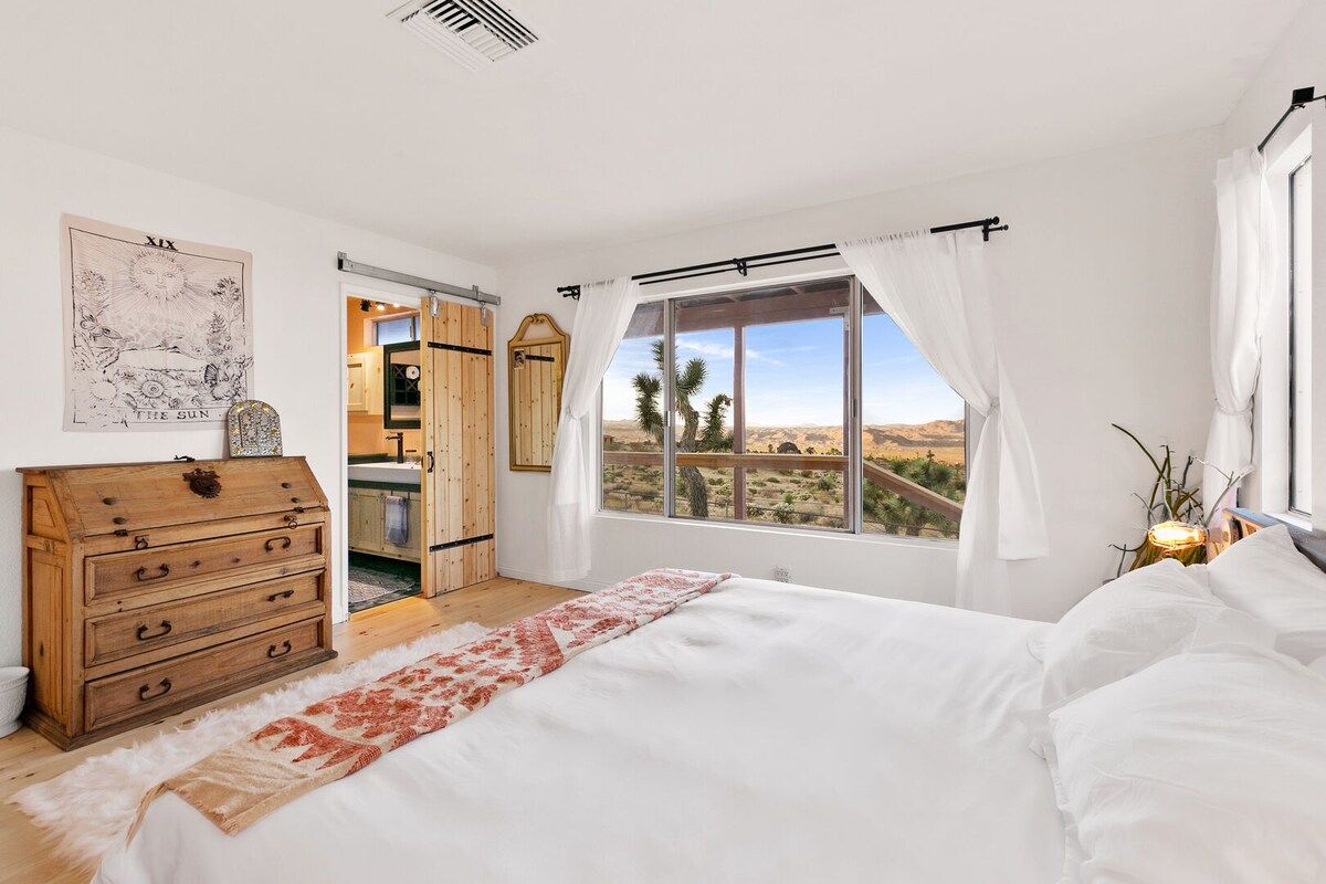 Ein Schlafzimmer mit einem großen Bett, einer Holzkommode, einem Fenster mit Blick auf eine Wüstenlandschaft und einer Tür, die in eine Küche führt.