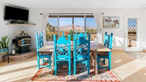 A dining area with a table and chairs, a television mounted on the wall, and a view of the mountains through large windows. - High View Haven - 5min to Park, Hot Tub, Fire Pit, BBQ (Joshua Tree)