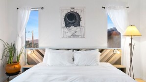 A hotel room with a bed, a wall-mounted poster, a potted plant, a standing lamp, and a view of the outside. - High View Haven - 5min to Park, Hot Tub, Fire Pit, BBQ (Joshua Tree)