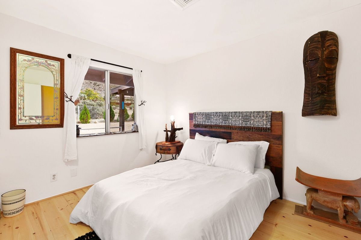 A bedroom with a bed, a window with curtains, a nightstand, a wall-mounted headboard, and a wooden sculpture.