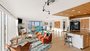 A spacious living area with a large window, a wooden ceiling, a kitchen with a black refrigerator, and a seating area with a colorful rug. - High View Haven - 5min to Park, Hot Tub, Fire Pit, BBQ (Joshua Tree)