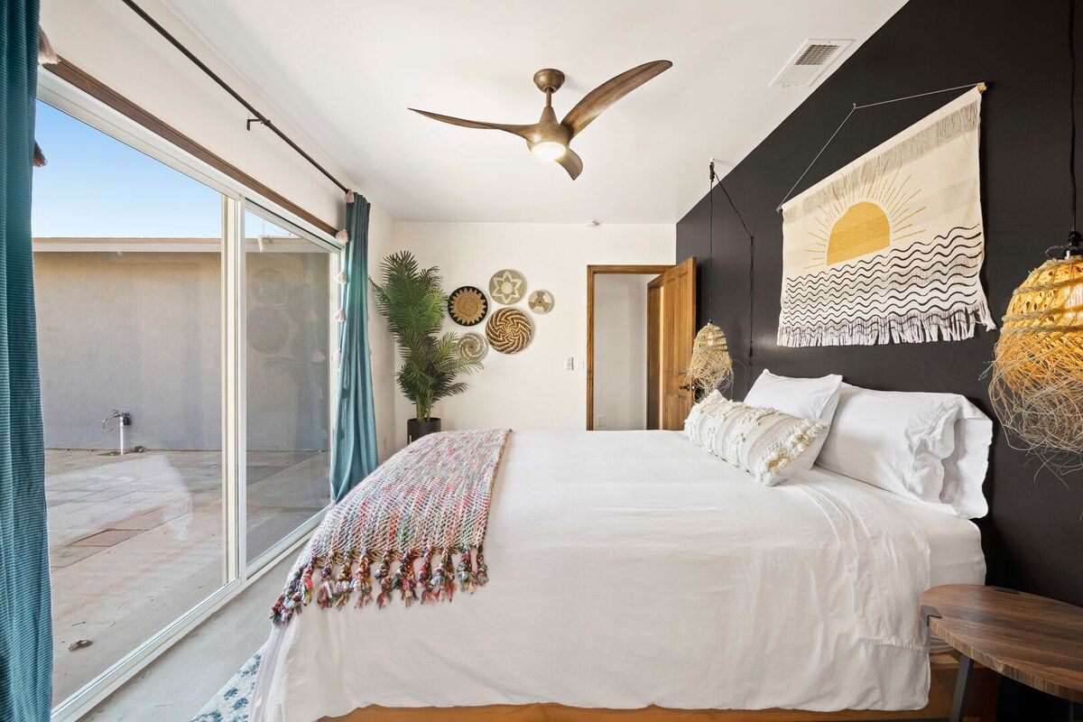 A bedroom with a bed, a ceiling fan, a large window, and a wall with decorative items.
