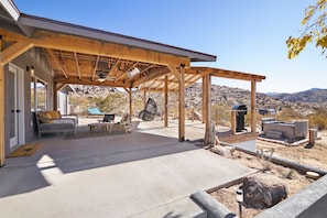 A covered patio area with outdoor seating, a hot tub, and a view of the surrounding desert landscape. - Dreamspace - Hot Tub, Fire Pit & MInutes to Park! (Joshua Tree)