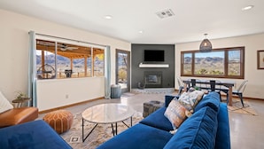 A spacious living room with a large sofa, a round coffee table, and a fireplace. There are large windows offering a view of the mountains. - Dreamspace - Hot Tub, Fire Pit, 5 Min to Park! (Joshua Tree)