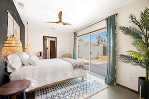 A bedroom with a bed, a ceiling fan, a large window with curtains, a view of the outdoors, and a potted plant.