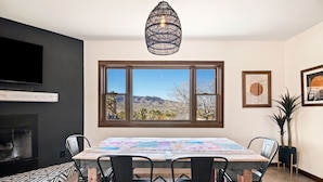 A dining area with a table, chairs, a fireplace, and a large window with a desert view. - Dreamspace - Hot Tub, Fire Pit, 5 Min to Park! (Joshua Tree)