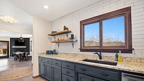 A modern kitchen with dark cabinets, a granite countertop, and a large window. - Dreamspace - Hot Tub, Fire Pit, 5 Min to Park! (Joshua Tree)
