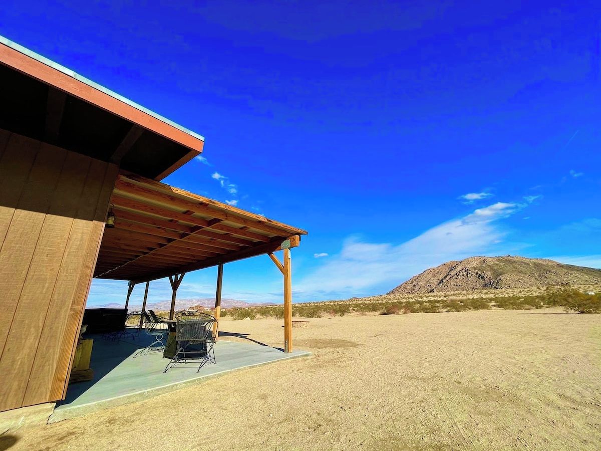 A covered outdoor area with a wooden structure, a grill, and a view of a desert landscape with a hill in the background.