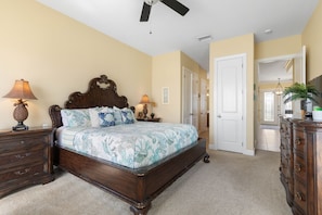 Quarto com cama, mesas de cabeceira, cômoda, ventilador de teto e porta que dá para outro espaço.