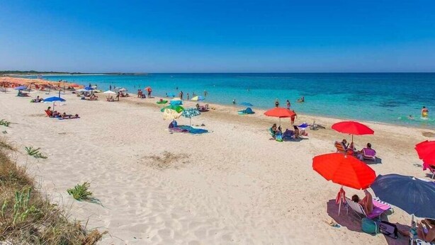 Een strand met mensen die ontspannen onder kleurrijke parasols, een heldere blauwe lucht en een kalme zee.