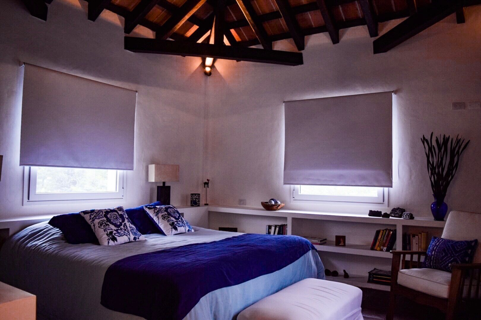 A bedroom with a bed, two windows with blinds, a nightstand, a chair, and a bookshelf.