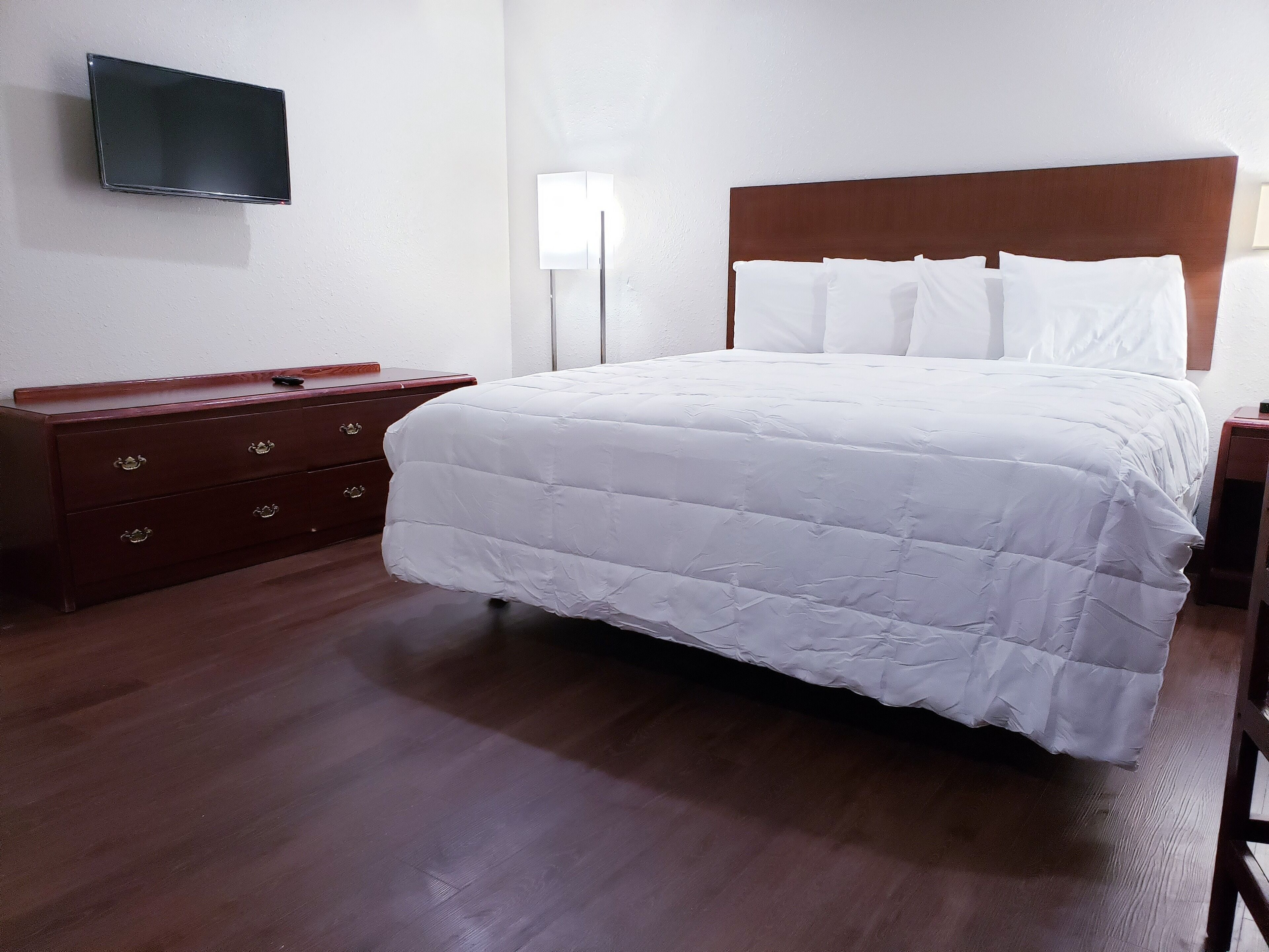 Een netjes opgemaakt bed met wit beddengoed, een houten dressoir met lades, een flatscreen televisie aan de muur bevestigd, en een nachtkastje met een lamp.