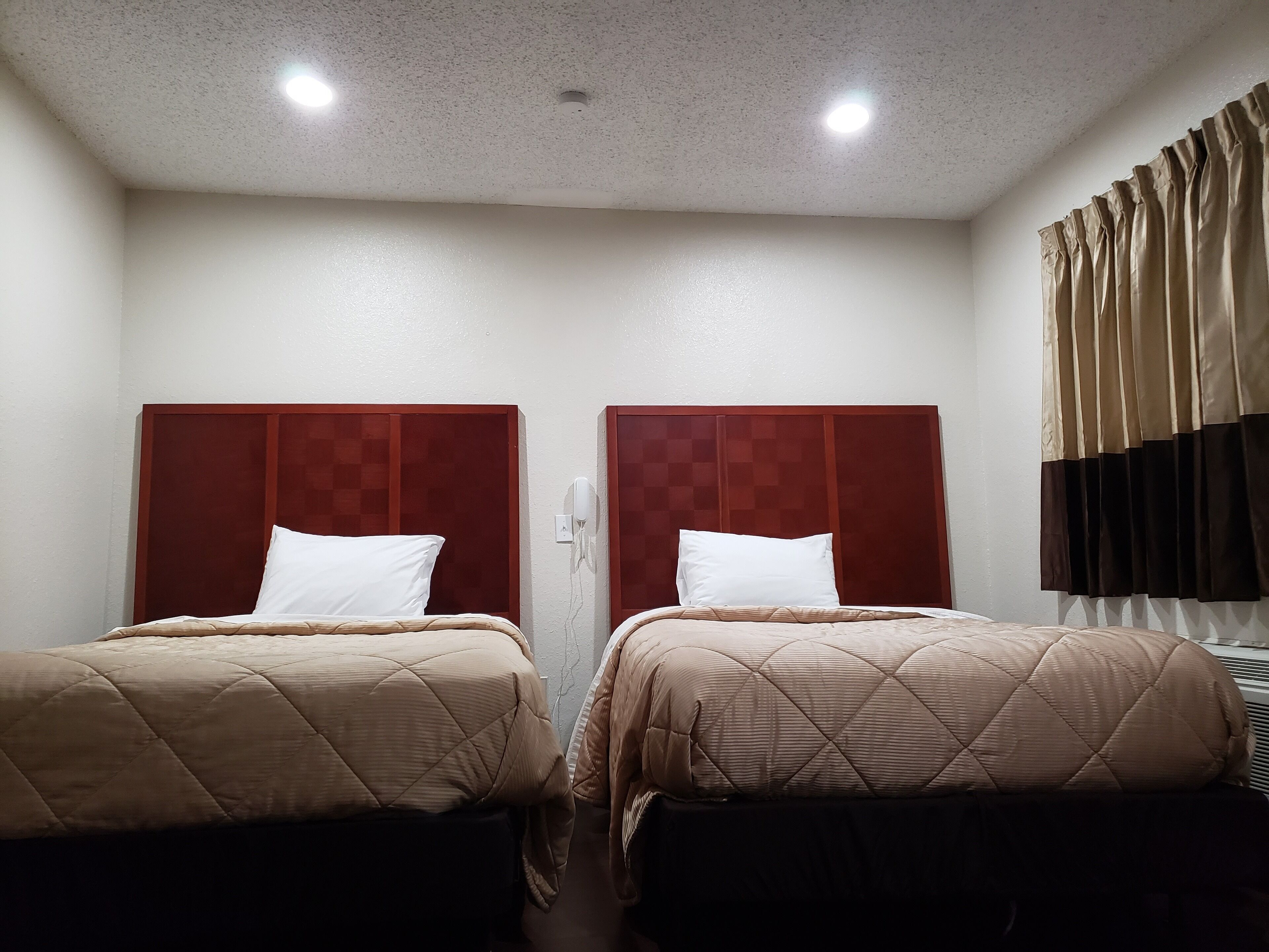 A hotel room with two beds, each with a wooden headboard and white pillows. There is a ceiling light and a wall-mounted air conditioning unit.
