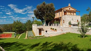 A two-story stone house with a balcony, a tennis court, and a large tree in the front yard. - Luxury Villa Retreat with Infinity Pool (Labin)
