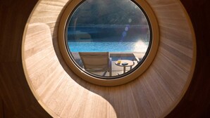 A round window with a view of a pool and lounge chairs.