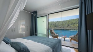 A bedroom with a large bed, a balcony with wicker chairs, and a view of a lake and hills.