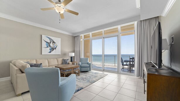 A spacious living room with a ceiling fan, a sofa, a blue armchair, a wooden coffee table, and a view of the ocean through large windows. - Phoenix Orange Beach II 704 by Liquid Life (Orange Beach)