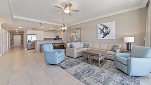 A spacious living room with a large sofa, two armchairs, a coffee table, and a chandelier. A ceiling fan and a kitchen are visible in the background. - Phoenix Orange Beach II 704 by Liquid Life (Orange Beach)
