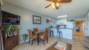 A spacious living area with a dining table, chairs, a television, and a ceiling fan. - Royal Palms 1002 by Liquid Life (Gulf Shores)