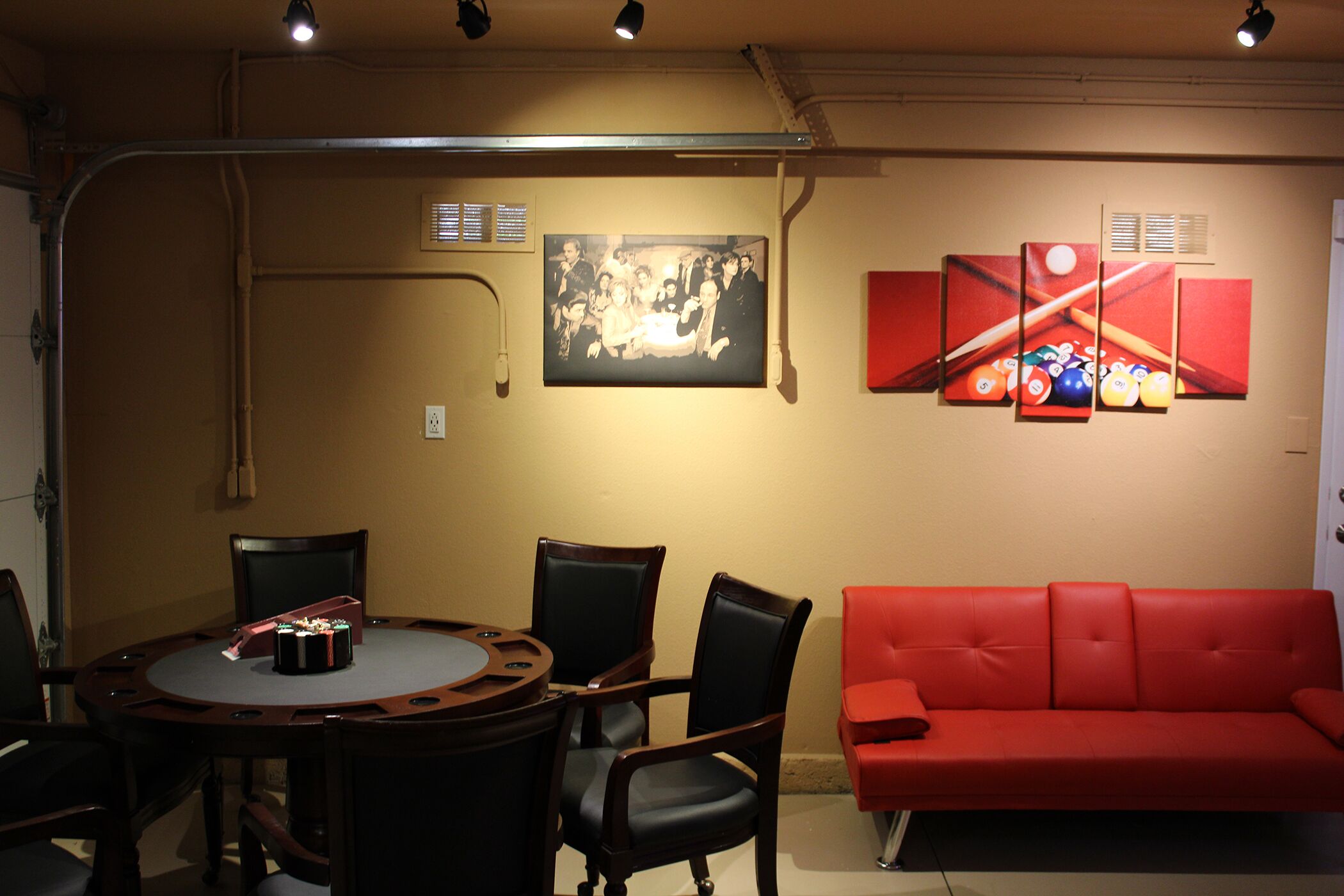 A room with a round table, four chairs, a red sofa, and wall art including a photograph and a pool table illustration.