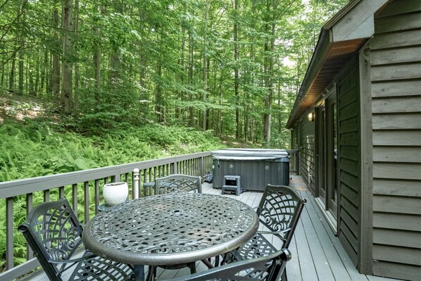 A wooden deck with a round metal table and chairs, surrounded by lush greenery and a hot tub.