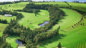 Vue aérienne d’un parcours de golf avec un club-house, des obstacles d’eau et des zones boisées environnantes.