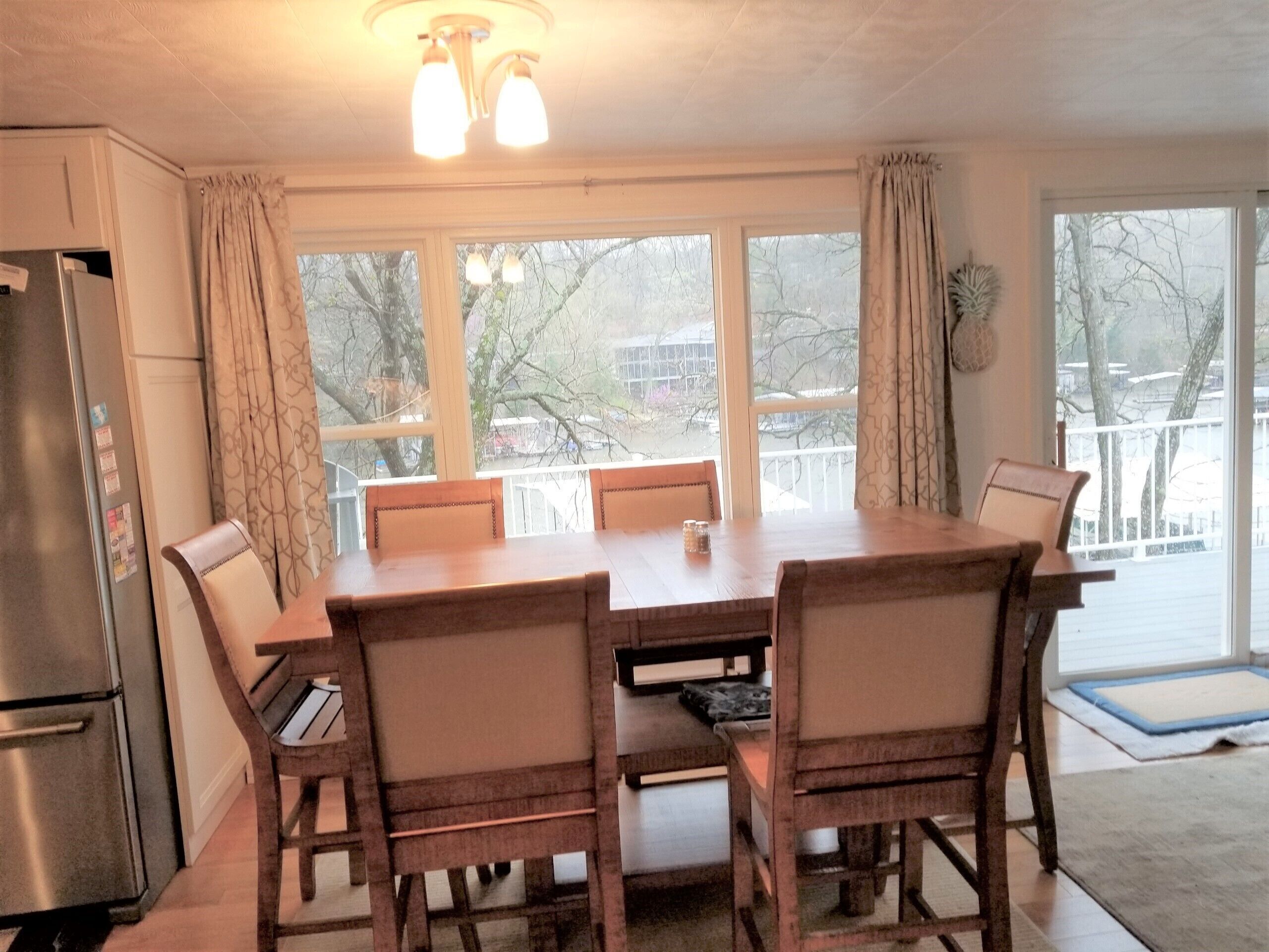 A dining area with a wooden table and chairs, a refrigerator, and large windows with curtains.