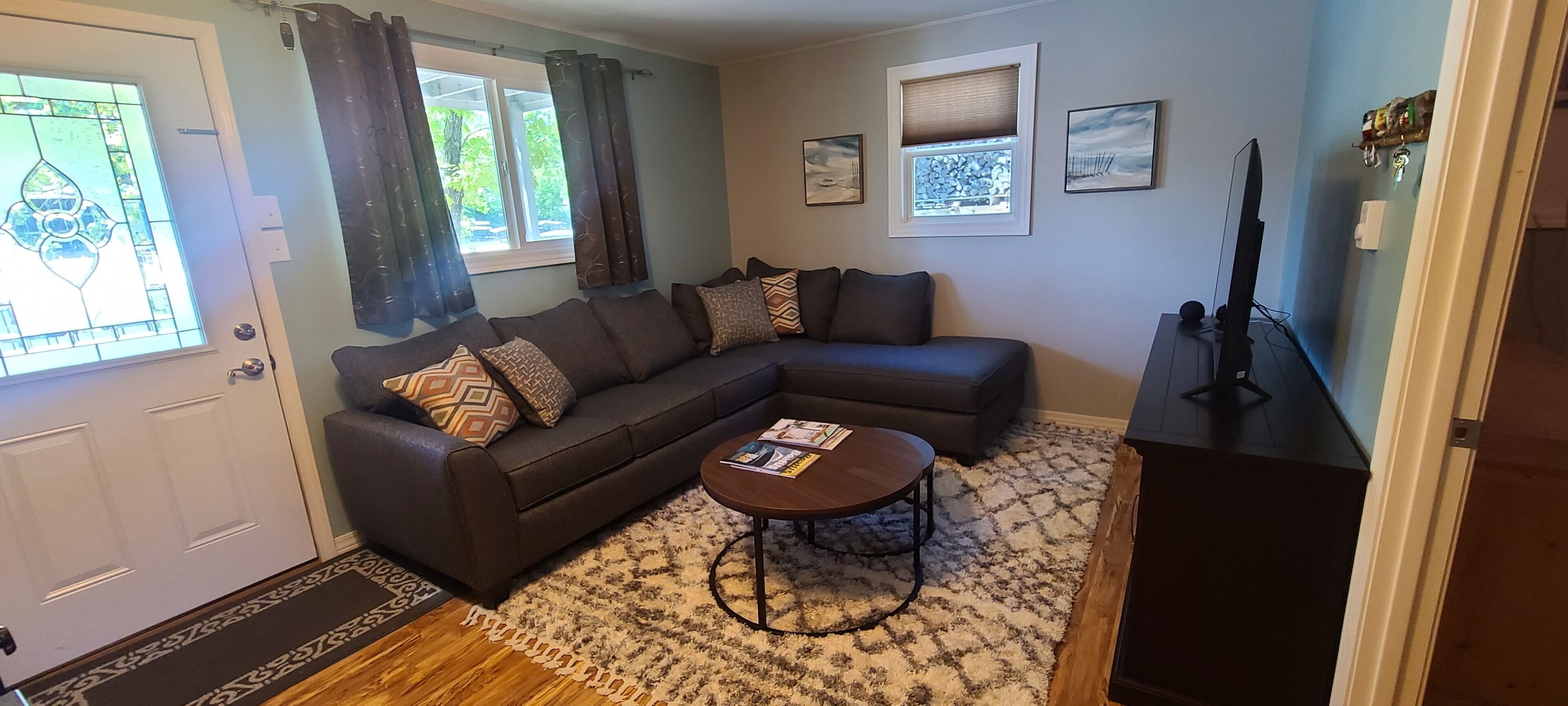 A living room with a dark brown sectional sofa, a round coffee table, and a patterned rug. There are two windows with curtains, a door, and a wall-mounted TV.
