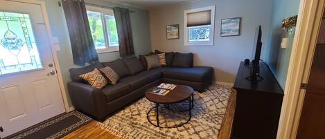 A living room with a dark brown sectional sofa, a round coffee table, and a patterned rug. There are two windows with curtains, a door, and a wall-mounted TV.