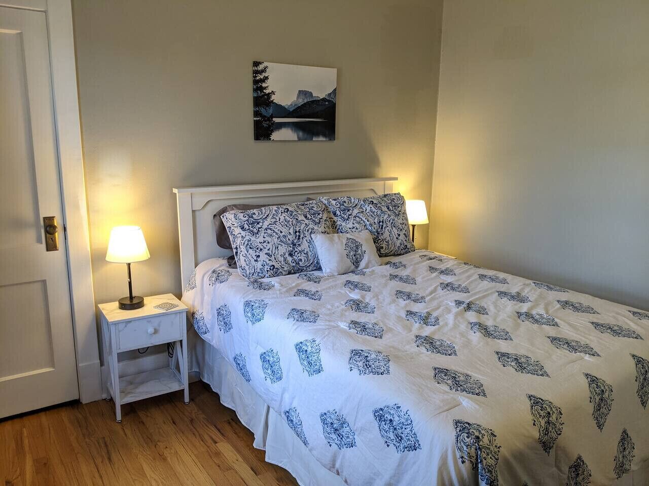 Een net opgemaakt bed met beddengoed met een patroon, een nachtkastje met een lamp en een ingelijst foto aan de muur.