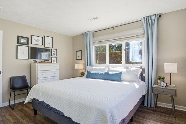 Un dormitorio con una cama grande, un aparador, una silla, una mesita de noche con lámpara y una ventana con cortinas.