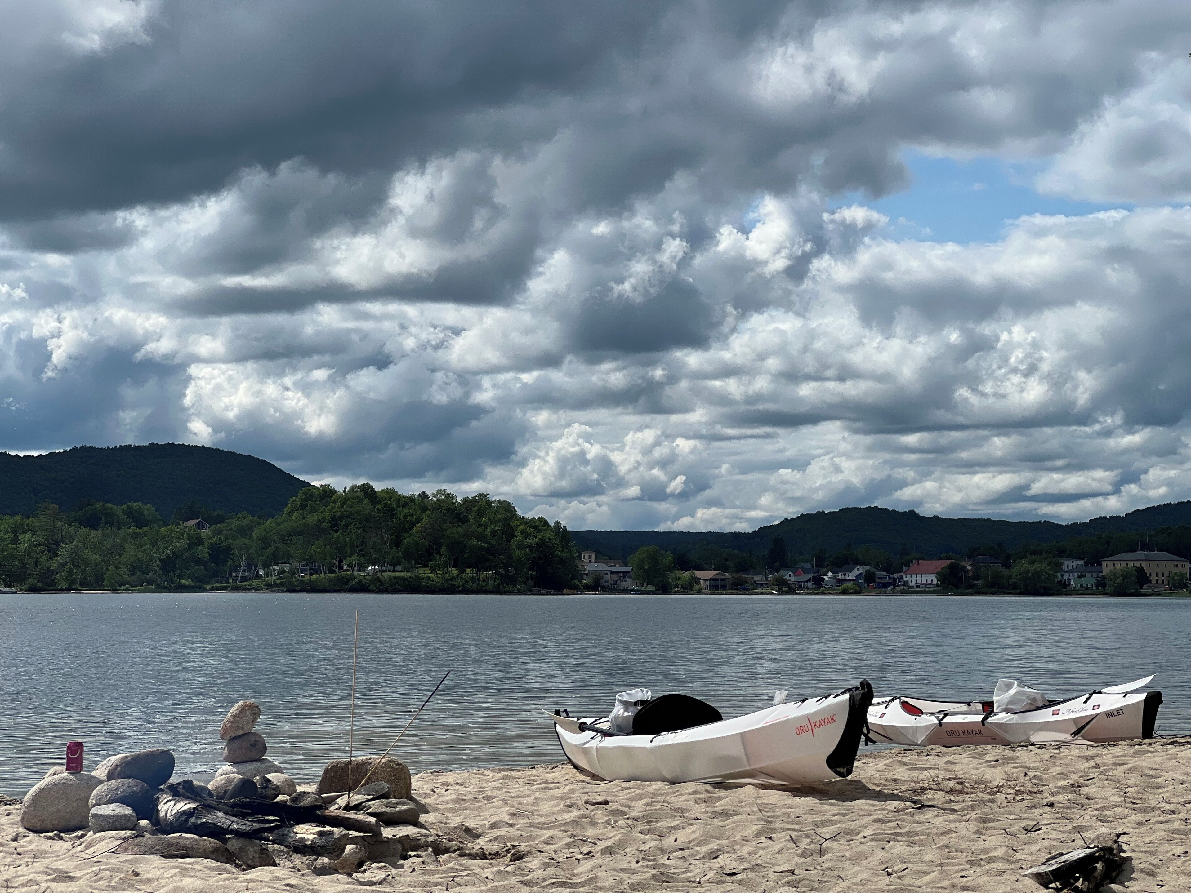 A lakeside view with kayaks and fishing rods on the shore, surrounded by hills and a cloudy sky.