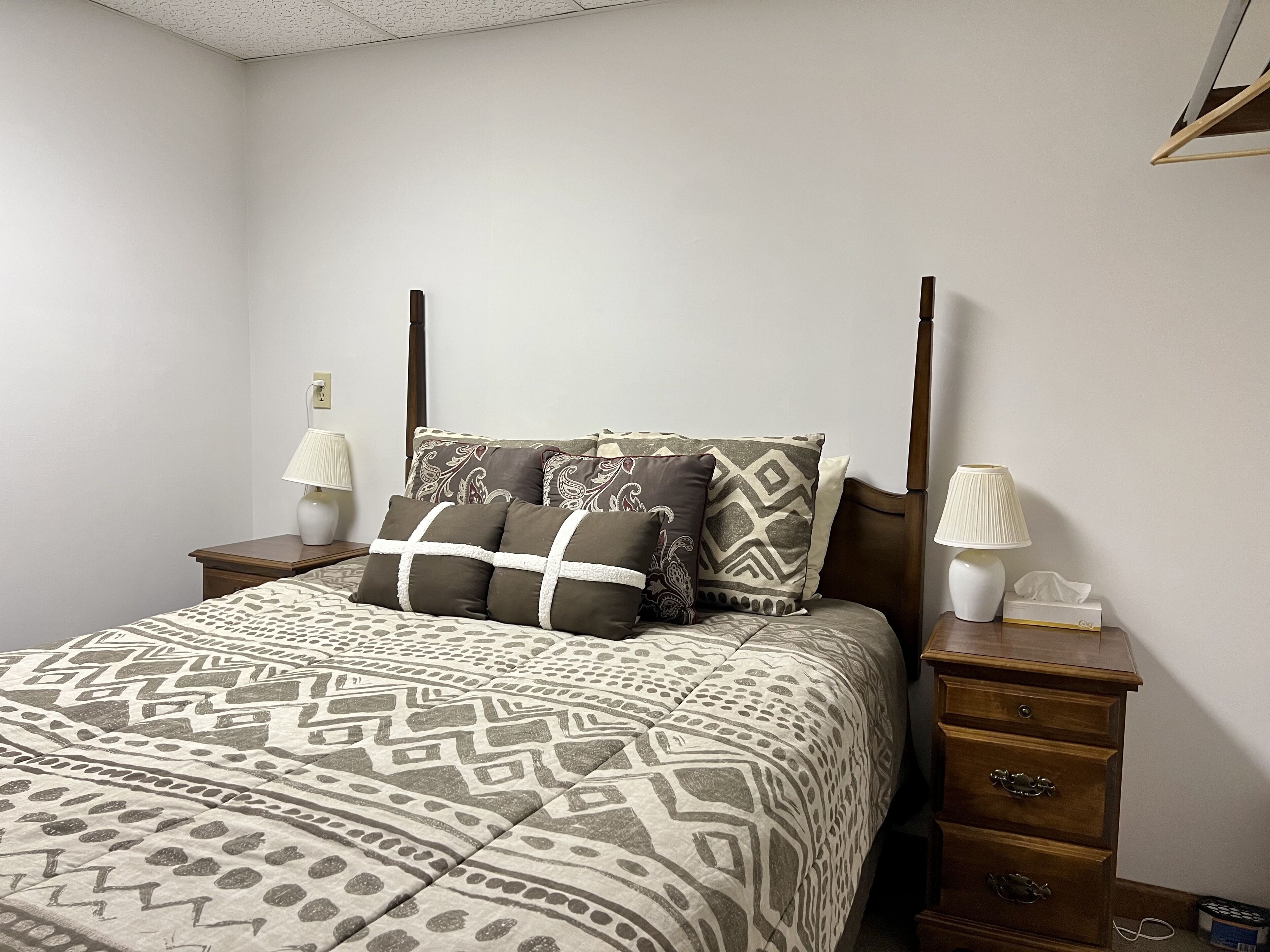A bedroom with a four-poster bed, patterned bedding, two bedside lamps, a nightstand with a tissue box, and a wooden dresser with drawers.